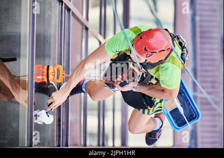 Arbeiter beim Bergsteigen waschen Glasfenster des Hochhauses, hängen an einem Kletterseil. Man Fensterputzer in Schutzhelm Reinigung Wolkenkratzer Fassade. Draufsicht. Stockfoto