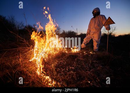 Feuerwehrmann Ökologe arbeitet auf dem Feld mit Wildfeuer in der Nacht. Mann in Anzug und Gasmaske in der Nähe brennendes Gras mit Rauch, mit Warnschild mit Schädel und Kreuzknochen. Konzept für Naturkatastrophen. Stockfoto