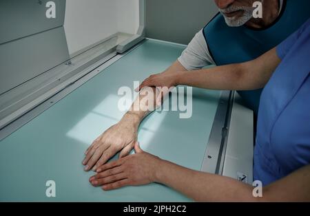 Radiologe, der die Hand eines männlichen Patienten auf einem modernen Röntgengerät Röntgen und die Hand mit Lichtstrahl an die richtige Stelle auf dem Röntgentisch legen kann Stockfoto