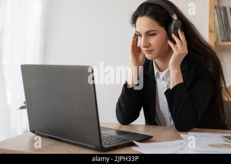 Seriöse Geschäftsfrau mit Kopfhörern, die sich ein Webinar auf dem Laptop ansieht, Computerkurse lernt, Anrufe tätigen, an Online-Konferenzen teilnehmen Stockfoto