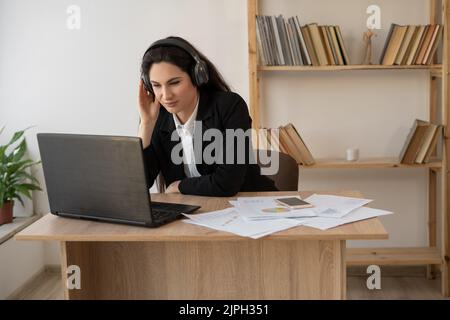 Seriöse Geschäftsfrau mit Kopfhörern, die sich ein Webinar auf dem Laptop ansieht, Computerkurse lernt, Anrufe tätigen, an Online-Konferenzen teilnehmen Stockfoto