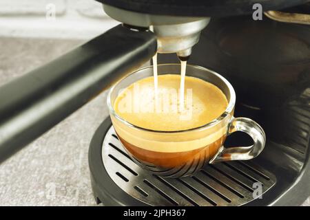 Glasbecher steht auf dem Gitter der schwarzen Kaffeemaschine und gießt Kaffee hinein. Prozess der Zubereitung von Kaffee. Stockfoto