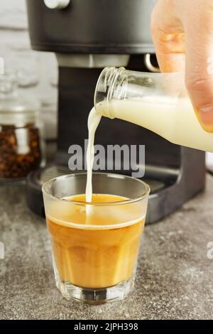 Barista gießt Milch in Cappuccino-Kaffeeglas auf graue Oberfläche mit Hintergrund der Kaffeemaschine Stockfoto