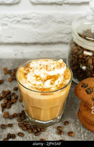 Karamell-Latte-Kaffee in einem Glas mit Schlagsahne und Schokoladenkekse. Kaffeebohnen auf grauer und weißer Ziegelwand mit Kopierfläche. Stockfoto
