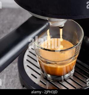 Glasbecher steht auf dem Gitter der schwarzen Kaffeemaschine und gießt Kaffee hinein. Prozess der Zubereitung von Kaffee. Stockfoto