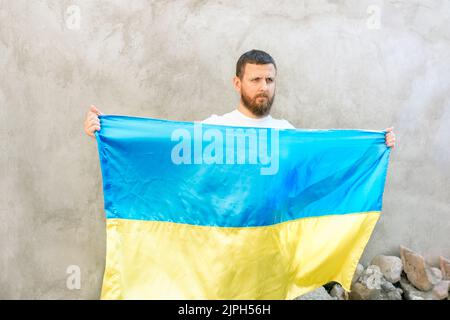 Der Mann hält die blau-gelbe Nationalflagge der Ukraine vor sich. Ein Kerl mit einem Bart in einem weißen T-Shirt und Jeans. Sonniger Tag. Verfassung und Unabhängigkeitstag der Ukraine. Weicher, selektiver Fokus Stockfoto