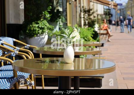 Straßencafé in der Stadt mit leeren Tischen im Freien im Hintergrund von Wanderern. Vasen aus Rosenblumen auf runden Tischen und gemütlichen Stühlen an sonnigen Tagen Stockfoto