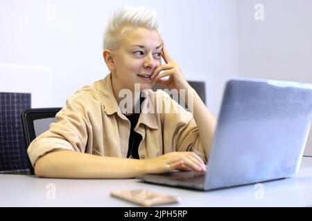 Mädchen mit schlauem Lächeln und kurzen blonden Haaren sitzen am Bürotisch mit Laptop. Tomboy Lifestyle, Konzept der Inspiration bei der Arbeit und Kreativität Stockfoto