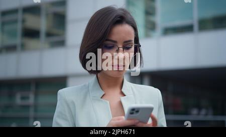 Hübsche junge Geschäftsfrau, die auf dem Smartphone chattet, gute Nachrichten liest, lächelt, während sie auf der Straße steht Stockfoto