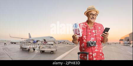 Reifer, männlicher Tourist, der einen Pass mit einem Flugticket hält und ein Smartphone auf dem Vorfeld eines Flughafens anschaut Stockfoto