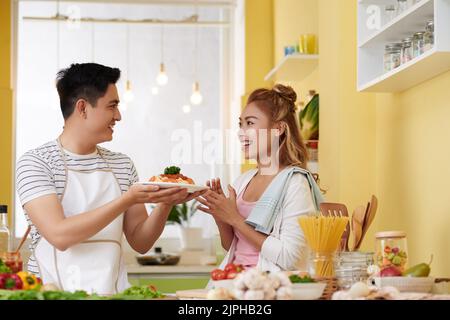 Asiatischer junger Mann, der seiner glücklichen Freundin einen Teller mit Spaghetti gibt Stockfoto