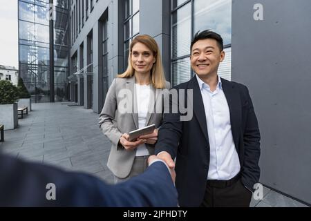 Business Meet, drei Kollegen begrüßen sich, Männer schütteln die Hände, Mitarbeiter vor dem Bürogebäude in Geschäftskleidung Stockfoto