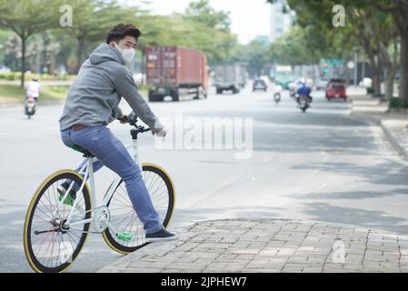 Junger asiatischer Mann mit Gesichtsmaske beim Radfahren in verschmutzter Stadt Stockfoto