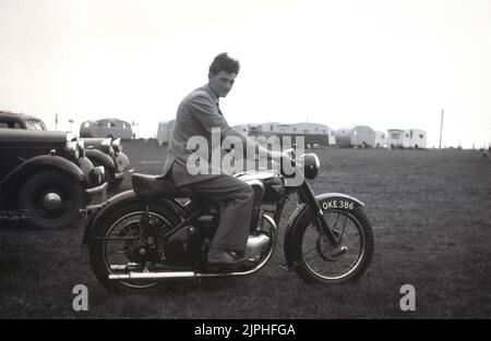 1950s, historisch, ein junger Herr, der auf einem britischen BSA-Motorrad auf einem Feld auf einem Caravanplatz sitzt, England, Großbritannien. Autos der Ära parkten. Gegründet 1861 als Birmingham Small Arms Company Ltd, für die Herstellung von Schusswaffen, wurde 1903 eine Motorradsparte der BSA gegründet und das erste Motorrad, das 1910 auf den Markt kam. Im Jahr 1950s war BSA Motorcycles der größte Motorradhersteller der Welt. Stockfoto