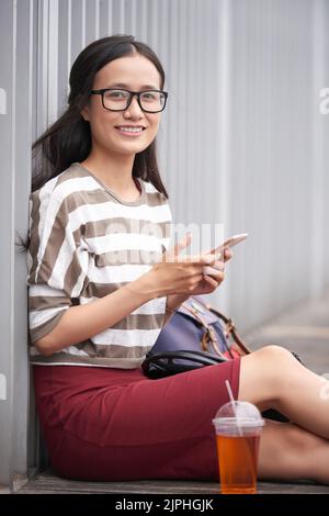 Porträtaufnahme einer attraktiven asiatischen Frau mit Brillen, die mit einem charmanten Lächeln auf die Kamera blickt, während sie auf dem Holzboden der Veranda sitzt und auf dem Smartphone im Internet surft Stockfoto