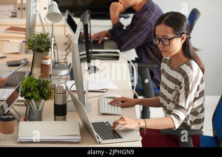 Arbeitsprozess im modernen Großraumbüro: Ziemlich junger Manager in der Freizeitkleidung, der statistische Daten analysiert und dabei moderne Computer- und Laptop-Profile verwendet Stockfoto