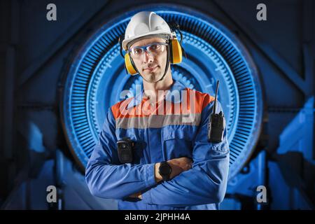 Porträt eines jungen professionellen Ingenieurs, der neben einem Gasturbinenmotor in einem Schutzhelm, Kopfhörern und einer Brille steht. Im Hintergrund ist ein Stockfoto