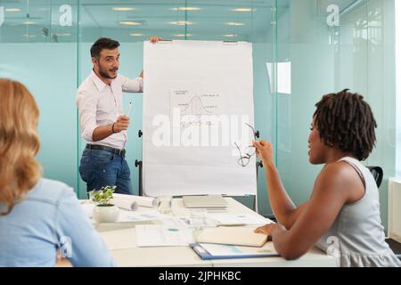 Gutaussehende bärtige Managerin in Casualwear, die am Markierungstafel steht und Ideen mit weiblichen Kollegen teilt, Inneneinrichtung eines modernen Sitzungssaals mit Glaswänden im Hintergrund Stockfoto