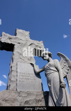 Ein steinerner Engel steht mit der Hand am Kreuz und ihre Flügel teilweise mit einem Blick von Traurigkeit und Verlust gegen den dunkelblauen Himmel ausgebreitet Stockfoto