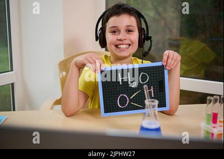 Fröhlicher Schuljunge, der dem Lehrer während des Online-Chemieunterrichts eine Tafel mit der chemischen Formel von Wasser H2O zeigt Stockfoto