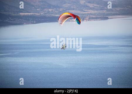 Ein Paramotor-Pilot, der an einem frühen Sommermorgen über der Mündung des Conwy River in Nordwales mit klarem blauen Himmel und einer unglaublichen Aussicht fliegt Stockfoto