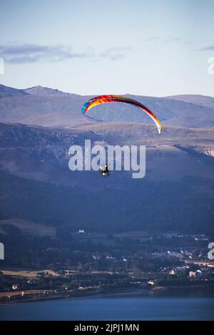 Ein Paramotorpilot, der an einem frühen Sommermorgen über die Mündung des Conwy in Nordwales fliegt, mit Penmaenmawr und walisischen Hügeln im Hintergrund Stockfoto