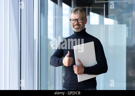 Porträt eines erfolgreichen, erfahrenen und reifen Geschäftsmannes, eines älteren grauhaarigen Mannes, der im Bürogebäude arbeitet, eines Finanziers, der lächelt und mit dem Laptop den Daumen nach oben zeigt Stockfoto