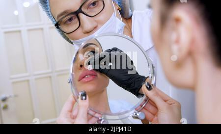 Arztpraxis, schöne Frau, die in den Spiegel schaut, wischt Arzt die Lippen des Patienten mit einer sterilen Serviette, bevor Injektionen von Hyaluronsäure in die Lippen, reinigt die Oberfläche mit einem Antiseptikum. Hochwertige Fotos Stockfoto