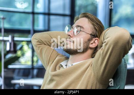 Mann, der sich zu Hause ausruhte, junger Mann mit geschlossenen Augen in einer Brille, Hände hinter dem Kopf, der tagsüber nippte, Freiberufler, der zu Hause aus der Ferne arbeitete Stockfoto