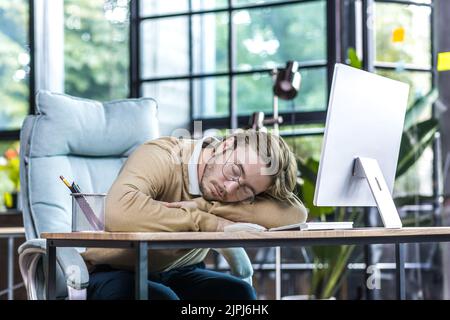 Junger Geschäftsmann ruht sich im Büro aus, Mann in Brille schläft auf dem Tisch, Freiberufler am Computer, Tagesschlaf Stockfoto