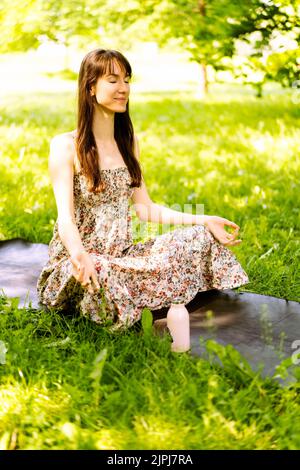 Eine schöne junge Frau sitzt auf einer Yogamatte im Park und macht Yoga oder Meditation. Speicherplatz kopieren. Das Konzept Fitness und ein gesunder Lebensstil. Stockfoto