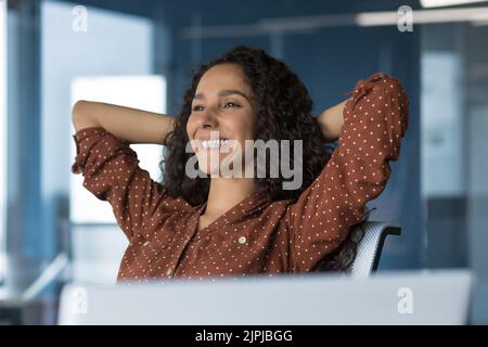 Schöne lateinamerikanische Geschäftsfrau, die sich im Büro ausruhte, Geschäftsfrau mit den Händen hinter dem Kopf, zufrieden mit dem Arbeitsergebnis, mit einem Laptop, einem Nahaufnahme-Foto Stockfoto
