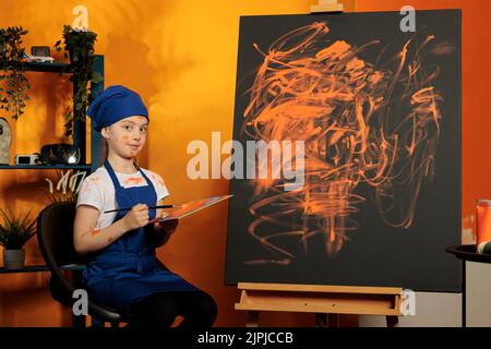 Portrait des jungen Künstlers Malerei Meisterwerk mit oranger Farbe aus Mischtablett und Farbpalette, Schaffung von Kunst-Design auf Leinwand mit Staffelei. Kreative Fähigkeiten, farbenfrohe Kunstwerke mit Aquarell zu malen. Stockfoto