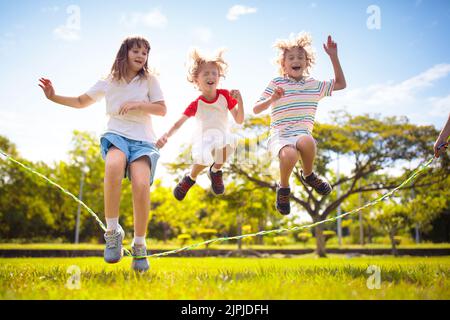 Glückliche Kinder spielen draußen. Kinder springen Seil im sonnigen Garten. Sommerurlaub Spaß. Gruppe von Schulkindern, die auf dem Parkspielplatz spielen. Stockfoto