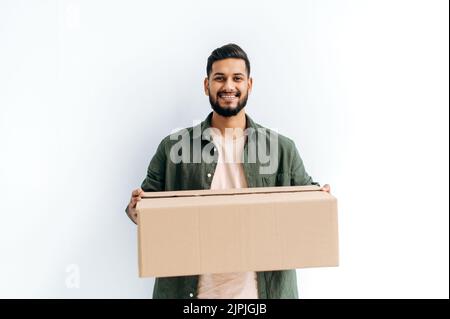 Positiv attraktiver, offen arabischer oder indischer junger Lieferer in legerer Kleidung, hält einen Karton, steht auf einem isolierten weißen Hintergrund, blickt auf die Kamera und lächelt freundlich. Lieferkonzept Stockfoto