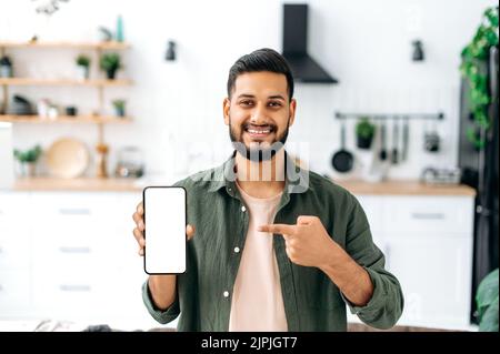 Modell des Telefonbildschirms. Lächelnder indischer oder arabischer junger Mann, der Smartphone mit leerem weißen Bildschirm, Kopierraum für Werbung oder Präsentation zeigt, im Wohnzimmer steht und auf die Kamera schaut Stockfoto