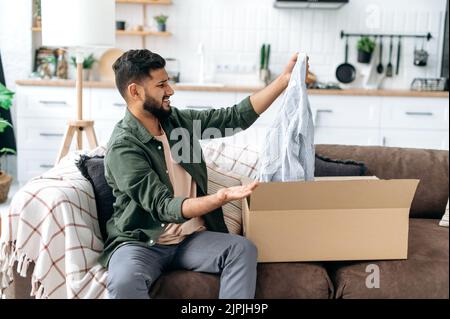 Frustrierter arabischer oder indischer Mann in legerer Kleidung, sitzt auf dem Sofa im Wohnzimmer, packt sein Paket aus, nimmt Kleider aus dem Karton, schaut sie enttäuscht und unzufrieden an Stockfoto