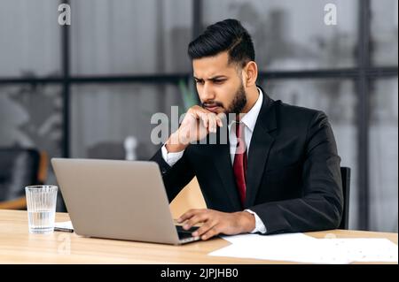 Verwirrt verwirrt verwirrt verwirrt ist der junge indische oder arabische Geschäftsmann, Unternehmer, in einem Anzug, sitzend in seinem modernen Büro, fokussierter Blick auf den Laptop-Bildschirm, unerwartete Nachrichten oder Nachrichten zu sehen, unter Stress Stockfoto