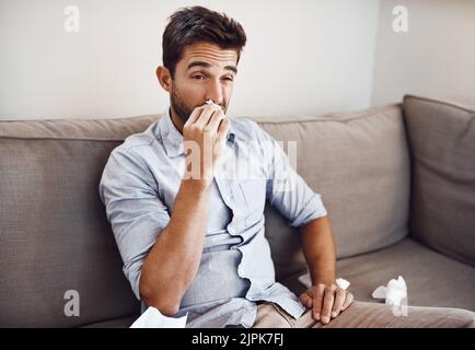 Eine verstopfte Nase zu haben ist so ärgerlich. Ein hübscher junger Mann bläst seine Nase mit einem Gewebe, während er sich auf einem Sofa zu Hause entspannt. Stockfoto