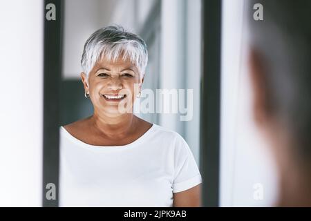 Nehmen Sie sich die Zeit, um Ihre Haut glänzen zu lassen. Eine fröhliche reife Frau, die ihre Spiegelung in einem Spiegel zu Hause betrachtet. Stockfoto