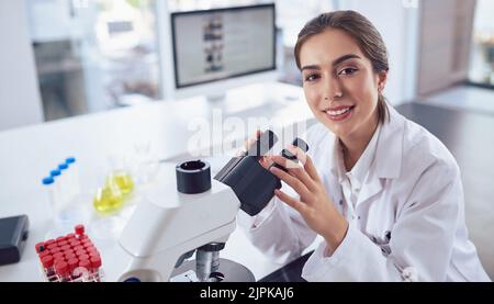 Wissenschaftler haben immer die modernste Ausrüstung. Porträt einer fröhlichen jungen Wissenschaftlerin, die ein Mikroskop benutzt, während sie die anschaut Stockfoto