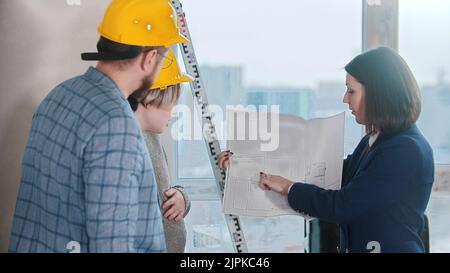Ein Immobilienmakler, der einem jungen Ehepaar in gelben Helmen ein Layout zeigt. Mittlerer Schuss Stockfoto