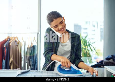 Ich bin nach dem Bügeln frei. Eine attraktive junge Frau benutzt ihr Handy, während sie das Bügeln zu Hause macht. Stockfoto