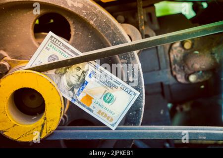 Ein 100-Dollar-Schein aus der Nähe vor dem Hintergrund des Motors eines ländlichen, hinter dem Traktor stehenden Traktors. Geld in der angetriebenen Riemenscheibe an den Riemen Stockfoto