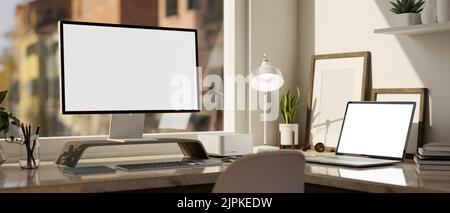 Moderner Büroarbeitsplatz mit Computer und Laptop, weißem Bildschirm, Tischlampe und Zubehör auf dem Tisch gegen das Fenster. Nahaufnahme. 3D Stockfoto
