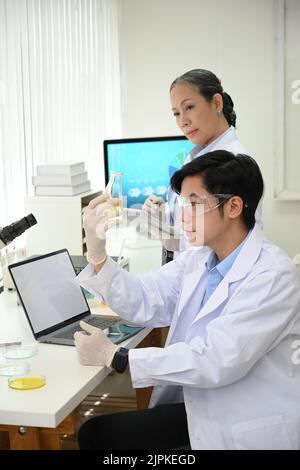 Ein talentierter junger asiatischer Wissenschaftler in einem weißen Mantel und einer weißen Brille, der einen Becher in der Hand hält und während seines Kollegen ein chemisches medizinisches Experiment im Labor macht Stockfoto