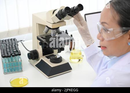 Eine professionelle Wissenschaftlerin im asiatischen Alter oder medizinische Chemikerin untersucht eine Virusprobe mit einem Mikroskop im Labor. Nahaufnahme Stockfoto