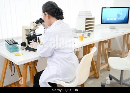 Eine erfahrene asiatische Wissenschaftlerin untersucht eine Virusprobe unter einem Mikroskop, während sie im Labor arbeitet. Stockfoto