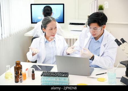 Eine professionelle Wissenschaftlerin im asiatischen Alter, die im Labor arbeitet, erklärt und ihrem jungen Wissenschaftler einen chemischen Rat gibt. Stockfoto