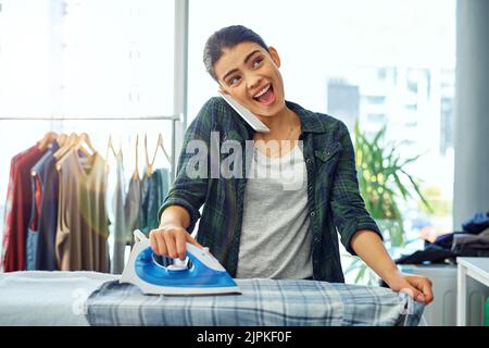 Ich bin gerade fertig mit dem Bügeln. Eine attraktive junge Frau, die ihr Handy benutzt, während sie das Bügeln zu Hause macht. Stockfoto